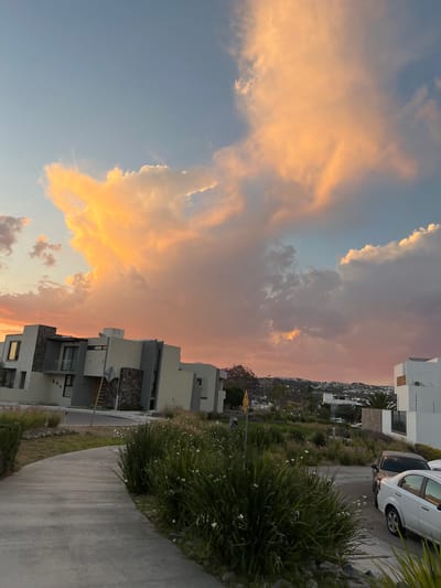The image shows a view in Arroyo Hondo, Mexico, at sunset. The sky is filled with clouds tinged with orange and pink hues from the setting sun. Modern houses line a street with well-maintained landscaping. A white car is parked near one of the houses. In the background, more houses and the hillside can be seen in the distance, fading into the twilight. The foreground features a sidewalk and vegetation.