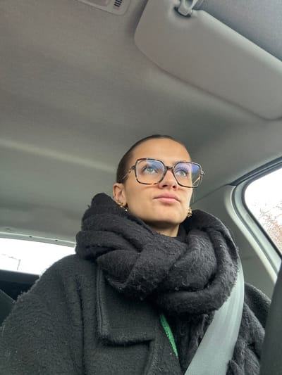 A female individual is depicted inside a vehicle in Saint-Cyprien, France. She occupies the front passenger seat, secured by a grey diagonal seatbelt. Her dark hair is pulled back, and she wears large, square-framed, tortoise-shell pattern glasses. Her light-colored eyes are directed upwards and slightly right. Attire consists of a textured black coat, a thick black knitted scarf, and a visible bright green undergarment. Multiple gold-colored earrings are present in both ears. The car's interior features a light grey fabric ceiling, a visible air vent, and a sun visor. A blurred outdoor environment is visible through the right-side window.