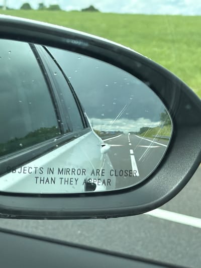 The image shows a side-view mirror of a car. The mirror reflects a highway stretching into the distance under a cloudy sky. The text "OBJECTS IN MIRROR ARE CLOSER THAN THEY APPEAR" is visible on the bottom of the mirror. The car is likely in motion on a road in a rural or suburban area, given the green fields visible in the background. There are no people or specific city landmarks identifiable in the image. The location of Пяршайскі сельскі Савет, Belarus is not evident from the visual information provided.