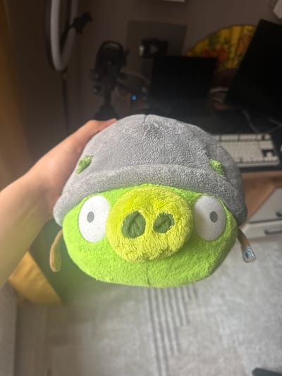A hand holds a plush toy of a green pig from the Angry Birds franchise. The pig has a grey, helmet-like head with a green patch on the side. Its face is bright green with large white eyes and a yellow snout with dark green nostrils. The pig has small brown ears. The background is blurred and appears to be an indoor setting with furniture and electronics, suggesting a home or office environment. The lighting suggests it might be daytime, but it's difficult to tell for sure due to the indoor setting and artificial lighting. There is no visible text in the image.