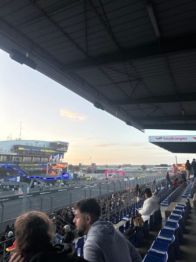 The image depicts a panoramic view from within a covered grandstand at a motorsport circuit, identified as the Circuit de la Sarthe in Le Mans, France, evidenced by the "AUTOMOBILE CLUB" building and French flags in the background, a primary identifier of the Automobile Club de l'Ouest (ACO). The scene is illuminated by the soft, warm light of either sunrise or sunset.
In the foreground, the dark grey, corrugated metal underside of the grandstand roof, supported by steel beams, occupies the top portion of the frame. Cylindrical black speakers are mounted beneath these beams. Below, the blue, hinged stadium seats of the grandstand are partially visible, with scattered spectators. A male with short dark hair and a beard, wearing a grey hooded sweatshirt, is prominent in the bottom center-right, looking towards the track. To his left, a person with long dark, curly hair is partially visible from behind. Further right, a person in a white puffy jacket is seated, looking down at a mobile phone.
The midground features the asphalt race track, bordered by a black safety fence and trackside barriers displaying sponsor logos, including "Samsung Galaxy A." Behind this, a dense crowd of spectators stands along the barrier. Beyond the track, to the left, a multi-story, modern building with "AUTOMOBILE CLUB" signage and French flags is visible, accompanied by a large digital screen displaying a close-up image of a racing car and other graphics. To the right, a large white sign with "TotalEnergies" in red is mounted on a metal structure, with several figures standing on a platform nearby. Distant buildings, trees, and a hazy horizon line are visible under a clear, pale sky with a single, elongated, reddish-orange cloud.