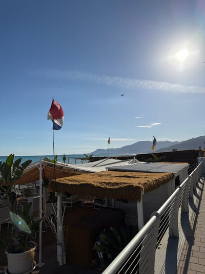 The image depicts a sunny outdoor scene at a beachside establishment in Vallecrosia al Mare, Italy. In the foreground, a modern metal railing lines a paved walkway. Behind the railing are several structures with thatched roofs, appearing to be part of a beach club or restaurant. Tropical plants, including large-leafed plants in white pots, are visible. A tall flagpole with a tri-colored flag (red, white, and blue) stands prominently on the left side of the frame. In the background, the turquoise sea meets a hazy coastline with mountains. The sky is clear blue with a bright sun casting lens flare and a few wispy clouds. Two birds are flying in the distance. The overall atmosphere suggests a warm, coastal environment.