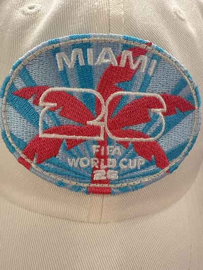 A close-up shot depicts a white baseball cap, featuring an oval-shaped embroidered patch. The patch's design incorporates the word "MIAMI" in white lettering along the left side. Centrally, stylized red palm tree graphics intersect with white outlined letters. The background consists of alternating light blue and white radiating stripes. To the right of the central design, the words "FIFA WORLD CUP" are embroidered in white. The cap's fabric appears textured, and the patch's stitching is visible. No people are visible in the image. The image is not linked to any specific location in Dania Beach, Florida.