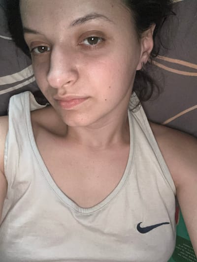 This media file shows a close-up, overhead shot of a young woman lying down, possibly in bed. She is looking towards the camera with a relaxed expression. The lighting suggests it could be nighttime or indoors with artificial lighting. She is wearing a light-colored tank top with a Nike logo on the chest. The background consists of striped bedding in muted tones of brown and gray. No specific location cues are visible beyond the implied domestic setting. The image is taken from a high angle, with the woman's face and upper body dominating the frame.