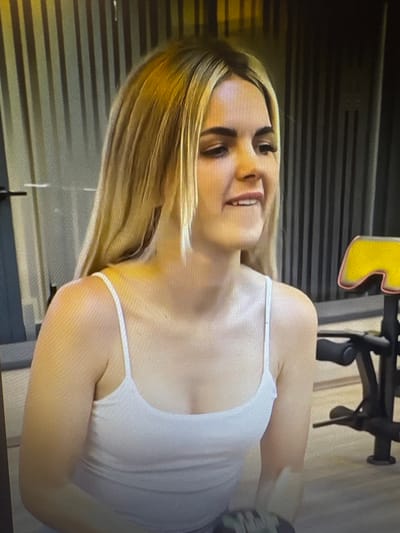 The media file shows a close-up, vertically oriented, full-body shot of a young woman with blonde hair. She is wearing a white tank top. She is smiling and appears to be looking towards the viewer, with her head tilted slightly. Her blonde hair is long and appears to be slightly messy, falling around her face and shoulders.

The background is a dark, horizontal striped pattern. To the left and slightly behind the woman, a yellow object with a rounded edge is visible, possibly part of a sign or logo. Behind the woman, a black metal structure is visible, which could be part of exercise equipment. The lighting appears to be artificial and somewhat dim. There are no clear indicators of the time of day or weather conditions. The emotion conveyed by the woman is one of happiness and perhaps amusement. The location is identified as Chorzów, Poland, but no specific landmarks or distinguishing features are visible in the image to confirm this.
