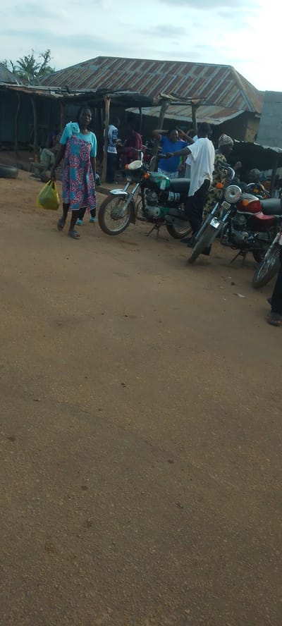 In Doka-Tofa, Nigeria, a group of people are gathered near a makeshift structure with a corrugated metal roof. The scene appears to be an informal market or roadside gathering. A woman in a blue and pink patterned dress walks from left to right, carrying a yellow plastic bag. Behind her, several other people are visible, some standing, some appearing to be interacting.
Two motorcycles are parked in the foreground. The one on the left is green with white and black accents, and its kickstand is down. The motorcycle on the right has a red fuel tank and appears to be a similar model. The ground is unpaved, consisting of packed dirt and small stones.
In the background, the structure has wooden posts and a tin roof that shows signs of age and rust. Some people are visible under the shelter, and the overall atmosphere suggests a busy, everyday scene. The sky is overcast, indicating either early morning or late afternoon, or possibly a cloudy day. There is no visible text in the image.
