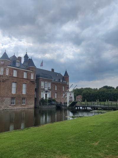 The image shows a historic brick castle situated on a moat in Isselburg, Germany. The castle has multiple towers and windows, and a drawbridge is visible, leading to a bridge lined with statues. A lush green lawn slopes towards the moat in the foreground, and a dense line of trees forms the background. The sky is overcast with dramatic clouds.