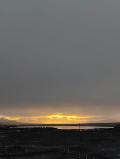 The image captures a panoramic view of a desolate landscape under a heavy, overcast sky. The upper portion of the image is dominated by a uniform, dark gray expanse of clouds. Towards the horizon, a band of intense yellow light breaks through the clouds, suggesting either sunrise or sunset. This bright light illuminates the clouds from below, creating a dramatic contrast with the otherwise somber sky.

Below the sky, a body of water, possibly a lake or a bay, stretches horizontally across the middle ground. The water appears calm and reflects the muted light from the sky. Along the distant shoreline, faint outlines of hills or mountains are visible, with a dusting of white suggesting snow or ice.

The foreground is a dark, uneven terrain, characterized by rough, uncultivated land. Dark shadows obscure details, but scattered piles of material and what appear to be construction stakes or poles are present, indicating some form of development or ongoing work in the area. A few indistinct structures, possibly machinery or vehicles, are faintly visible on the dark ground. Two utility poles with visible crossbars stand out against the horizon. The overall impression is one of a remote and rugged environment.

The location is not specified within the provided image data. Therefore, it is not possible to definitively state the location is in Mayskiy, Russia.