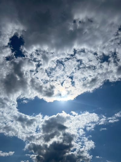 A vertical, low-angle shot captures a dynamic sky over Sobishchytsi, Ukraine, filled with dramatic, varied cloud formations. The bright sun peeks through a large, bright cloud mass in the upper center, casting rays and creating a halo effect. Below this, darker, more ominous clouds drift across a clear, vibrant blue sky. The clouds exhibit a range of textures, from wispy and thin to dense and shadowed, suggesting a mix of weather conditions. The image is devoid of any visible structures, people, or ground-level objects, focusing solely on the atmospheric phenomena.