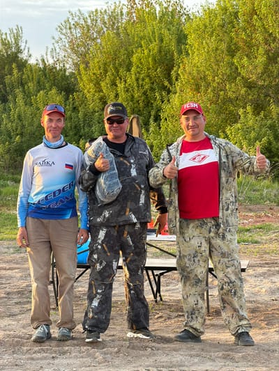 Three men are standing outdoors.
The first man, closest to the viewer, is wearing a long-sleeved, blue and white shirt, khaki pants, and a red baseball cap.
The second man is wearing a black jacket with grey accents and black pants. He is also wearing sunglasses and a black baseball cap. He is holding a bundled object wrapped in clear plastic.
The third man is wearing a camouflage jacket, a red t-shirt with a white logo, and a red baseball cap. He is giving a thumbs-up.
All three men are standing near a folding table.
The background is a combination of trees and a dirt ground.
Based on the "FCSM" on one of the caps, it is likely the location is in the city of Krasnyi Bor, Russia.