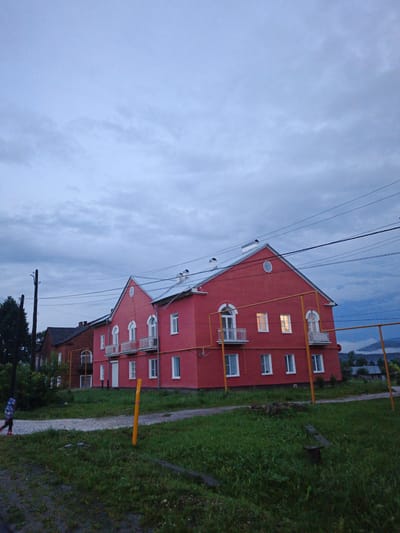 This media file depicts a residential building in Medvedyovka, Russia, during what appears to be dusk. The sky is overcast with heavy, dark clouds, suggesting imminent or ongoing precipitation. The main subject is a vibrant red, two-story building with a metal roof and several arched windows. White balconies with railings are visible on the upper floor. Some windows on the upper floor are lit, indicating occupancy.

In front of the building, there is a grassy area with a gravel path. A solitary child, wearing a blue and white striped garment and dark pants, walks away from the camera on the path. A tall wooden utility pole with wires is situated to the left of the building, and a yellow gas pipe runs along the front of the red structure. In the background, a darker brick building is partially visible, along with trees and a hint of a mountainous landscape under the gloomy sky. The overall mood is somber and quiet, characteristic of a rural setting at twilight. No visible text is present in the image.
