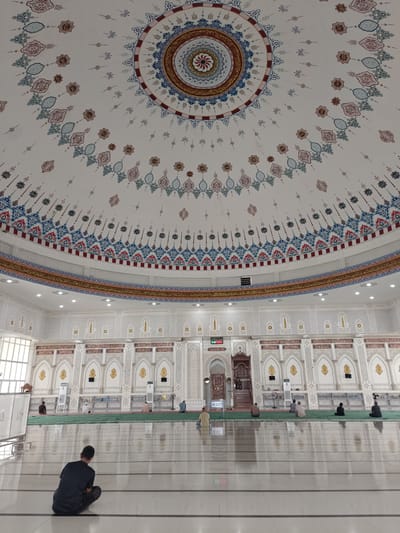 The image shows the interior of a mosque, possibly located in Paya Tringgadeng, Indonesia. The dome of the mosque is intricately decorated with geometric patterns and calligraphy in shades of blue, red, and gold. The walls are also adorned with similar ornate designs, featuring arched niches and Arabic inscriptions. Several individuals are seen performing prayers on green carpets laid out on the polished floor. In the foreground, a person is seated facing away from the camera. The environment appears to be well-lit with artificial lighting.