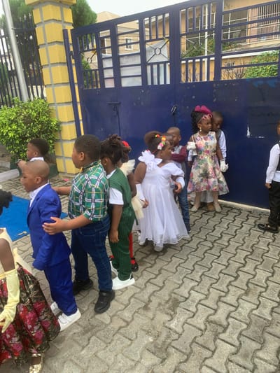 The image shows a group of children in a school uniform standing in a line outside a school gate in Close, Nigeria. The children are all neatly dressed. There are two girls in front, followed by a boy in a plaid shirt, and another boy in a suit. The children are all looking at the camera. The gate is made of blue metal and has a sign on it. There is a paved path leading up to the gate. The children are standing on the paved path. The environment is that of a school in Close, Nigeria. There is a patch of green bushes on the left and a yellow brick wall behind the group. The blue gate is on the right. The children are all interacting with each other and seem to be enjoying themselves. The scene is peaceful and pleasant.