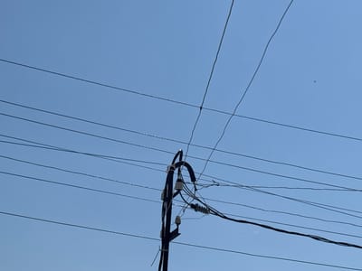 In Jrarat, Armenia, against a clear, bright blue sky, a power pole stands prominently in the frame. The pole, dark in color, is adorned with multiple insulators, both grey and white, from which a complex network of black electrical wires emanates. These wires stretch across the sky in various directions, some appearing taut and others with a slight sag. The clear sky suggests a daytime scene, possibly with the sun positioned to create subtle shadows or highlights on the wires, though they are not a dominant feature. The overall impression is one of urban infrastructure against a natural backdrop, with the intricate web of wires forming a significant visual element.