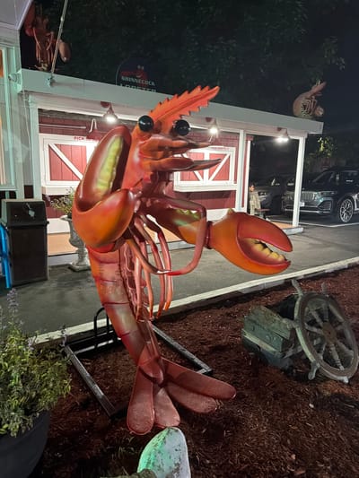 A large, bright orange lobster sculpture stands prominently in front of a building with red siding and white trim. The building has a sign that reads "SHINNECOCK LOBSTER." The lobster sculpture is detailed, with segmented body parts, claws, and antennae. In the foreground, there is a planter with green foliage and a wooden ship's wheel resting on a weathered wooden crate. The ground is covered in dark mulch. In the background, several cars are parked, and trees with dark foliage are visible, suggesting it is nighttime. The scene is outdoors, likely a restaurant or seafood establishment, located in the Town of Southampton, United States of America.