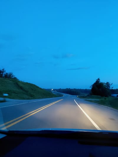 The image shows a road at dusk or twilight. The sky is a deep blue with some faint clouds. The road has two yellow lines in the center and a white line on the right side. Headlights from the viewpoint of the car illuminate the road ahead. To the left of the road, there is a grassy hill with some trees. To the right, there are more trees and what appears to be a body of water or a distant landscape. The scene appears to be a rural or suburban area. There are no people or animals visible. The location is not specified in the image, and therefore cannot be identified as being in the city of Jeffersonville, United States of America.