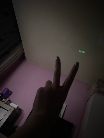 The image captures an indoor scene, likely in a room in Verkhnevesyoloye, Russia, characterized by dim lighting suggestive of nighttime. In the mid-right portion of the frame, a human hand is visible, making a two-finger "peace" or "victory" sign with fingers extended upwards. The fingernails of the visible fingers are painted with a dark blue or black polish.

On the light-colored ceiling in the upper left quadrant, a green digital display is projected. The primary numbers read "53:42". Above the "42" is a small "S", and above the "53" there is a less distinct symbol, possibly a "3" or "3°". This projection appears to be a clock, timer, or a temperature display.

The room features a wall painted a light purple or lavender color, visible on the right side of the image. In the lower left corner, a dark curtain or drapery can be seen, indicating the presence of a window. The bottom right area shows dark, indistinct furniture, possibly a shelf or dresser, with some vague objects placed on it. The overall ambiance is dark, with the projected green light being a focal point on the ceiling.