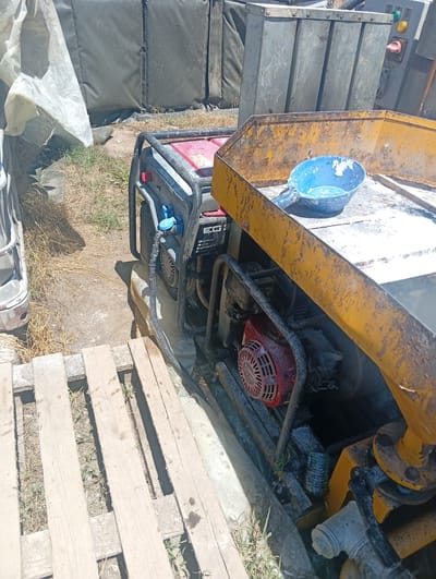 In Chelopech, Bulgaria, a red and black portable generator is positioned outdoors on a dirt and grass surface. The generator is connected to a black ribbed hose. Adjacent to the generator, to the right, is a yellow industrial piece of equipment with a blue scoop resting on its surface, the scoop containing a white powdery substance. In the background, a large, dark gray tarp is visible. To the left of the generator, a white tarp is draped over an unseen object. In the foreground, a wooden pallet lies on the ground. The scene appears to be an outdoor work or storage area.