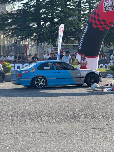 In Poti, Georgia, a blue and grey BMW E36 is parked on an asphalt road, possibly during a drift event, indicated by "GDS PARK DRIFT" decals on the car. The car is decorated with stickers, including a cartoon character on the rear quarter panel and "betlive RACING SCHOOL" on the rear bumper. A large inflatable arch with "betlive.com" printed on it stands behind the car, suggesting a sponsorship or event branding. A crowd of spectators is gathered behind a metal fence, observing the scene. The environment appears to be outdoors, with trees and a building visible in the background. A woman in a red skirt and white top, and another woman in a high-visibility vest, stand near the arch.