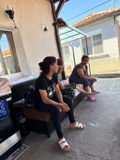 In Vinitsa, Bulgaria, three women are seated on a dark-colored sofa outside a house. The woman in the foreground, wearing a black t-shirt and black leggings, is looking to her right. Behind her, another woman with reddish-brown hair is also looking to her right. A third woman, wearing a dark purple patterned dress and pink slippers, is seated to the right and looking towards the viewer. The setting appears to be a courtyard with a concrete floor and a white-rendered wall. A chain-link fence is visible in the background, along with a residential building with a tiled roof. There are also some household items visible, including a black backpack on the sofa, a bottle of water, and some folded fabric on a surface to the left. The overall scene depicts a casual outdoor gathering.