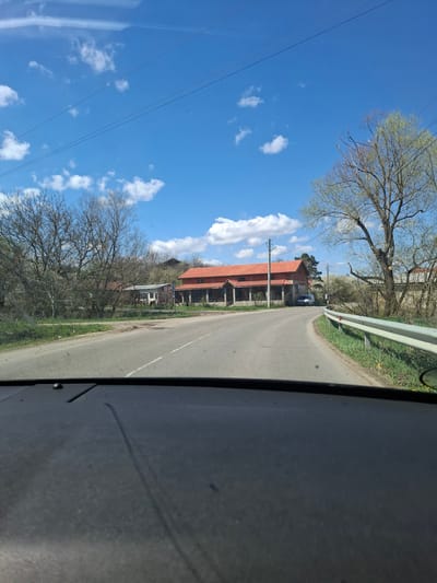 The image presents a view from inside a vehicle traveling on a rural road near Studeno Buche, Bulgaria. On the left, a clear blue sky with scattered white clouds is visible, punctuated by utility lines. Trees with bare branches line the roadside, interspersed with buildings having red-tiled roofs. The asphalt road stretches ahead, bordered by a metal guardrail on the right. The interior of the vehicle, including a portion of the dashboard or door panel, occupies the right side of the frame. A single vehicle is visible ahead.