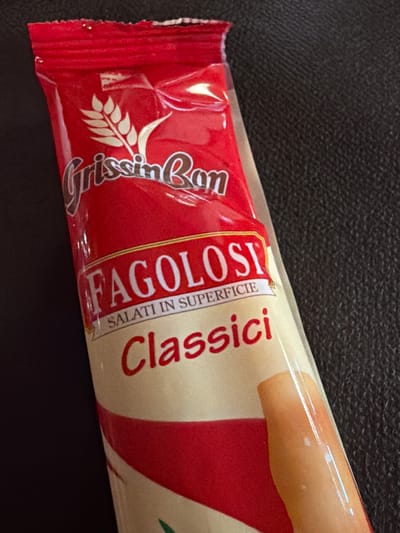 A close-up, high-angle shot shows a red and beige package of GrissinBon "Fagolosi Classici" breadsticks against a dark, textured surface, possibly leather. The packaging is sealed at the top with a crimped red seal. A golden wheat stalk is depicted on the upper part of the package, alongside the brand name "GrissinBon" in stylized brown lettering. Below this, the words "FAGOLOSI" are prominently displayed in large white letters with a red outline, followed by "SALATI IN SUPERFICIE" in smaller black text. The word "Classici" is written in a flowing red script. The bottom portion of the package reveals a portion of the breadsticks themselves, which appear golden brown and have a slightly shiny, oily sheen. The overall impression is that of a food product packaging. There is no indication of location within the city of Sauris / Zahre, Italy, as the image focuses solely on the product.