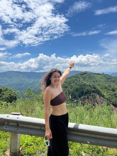 A young woman stands at a scenic overlook on a mountainside in Son Dien, Vietnam. She is wearing a brown strapless top and black pants, holding a pair of sunglasses in her right hand. Her left arm is raised, pointing towards the expansive, lush green landscape that stretches out before her. The background is dominated by rolling hills covered in dense vegetation, with a bright blue sky dotted with fluffy white clouds above. A metal guardrail is visible in the foreground, indicating the presence of a road or viewpoint. The overall scene suggests a tourist enjoying the natural beauty of the region.