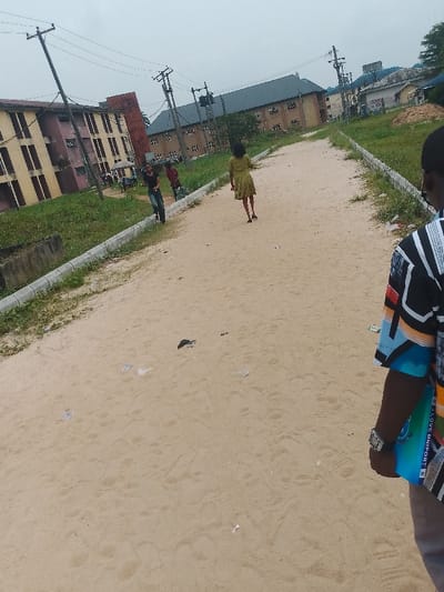 The image depicts an unpaved, sandy pathway in Choba, Nigeria, under an overcast sky. The pathway, showing numerous footprints and scattered litter (including white paper scraps and a dark object), is bordered by low concrete curbs. Beyond the curbs, green grass and some taller weeds are present.

In the foreground, on the right, the partial upper body of an individual is visible from the chest down, facing away from the viewer. This person wears a short-sleeved shirt with a multi-colored geometric pattern in black, white, blue, orange, and green. A metallic wristwatch is on their left wrist. They hold a light blue rectangular object, possibly a book or folder, which bears the vertical inscription "I LOVE UNIPORT". Light-colored trousers are visible below the shirt.

In the mid-ground, a female figure is seen from behind, walking away down the sandy path. She is wearing a light green, knee-length dress and dark footwear.

Further down the pathway, on the left side, two additional individuals are visible near the curb. One person, wearing a dark top and light-colored trousers, is partially facing left. Another individual in a red shirt is partially obscured behind them. Several other blurred figures are discernible closer to the buildings on the left.

The left side of the pathway is flanked by a multi-story building with yellow/beige walls and brown window shutters, adjoined by a section of a red-hued building. Multiple dark utility poles with numerous overhead lines are present on both sides of the pathway. In the far background, several other multi-story buildings, including one with a dark roof and another with a light blue roof, are visible amidst scattered trees.