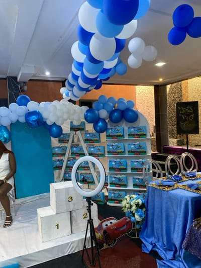 The image shows a party setup for a child's birthday in Bonadikombo, Cameroon. There are blue and white balloons in an arch, a blue backdrop, a cardboard stand with gift bags, and a small wooden stage with the numbers "1", "2", and "3" written on each step. A woman in a white dress is sitting in a clear plastic chair, and a ring light and tripod are on the floor to the right of the stage. A red toy car is on the floor. There are also several blue tablecloths on tables to the right. There are decorative items, such as flowers and small gold-colored decorative items. The location is in an indoor space with a tiled floor and a partially visible brown wall.