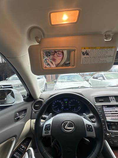 A car interior features a sun visor with a mirror reflecting a woman in glasses, puckering her lips, and holding a phone. The visor has a warning label. Adjacent is a dashboard with climate control buttons and a steering wheel with the Lexus logo. The speedometer reads 237. Outside, multiple cars are visible, suggesting the car is parked in a lot or on a street. The time displayed is 2:21.