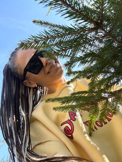 A woman with sunglasses is smiling upward, framed by evergreen branches against a bright blue sky. She is wearing a light yellow hooded sweatshirt with red lettering. Her hair is styled in braids with a mix of dark and light strands. The scene appears to be outdoors. Due to the limited information, the exact location within Yaroslavl', Russia cannot be determined.