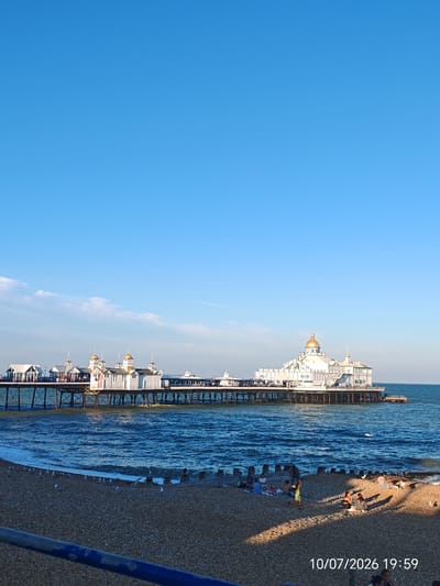 The image captures a wide shot of the Eastbourne Pier in Eastbourne, United Kingdom, on a sunny day. The pier, an iconic Victorian structure, extends into the bright blue sea, its white buildings and golden domes prominent against the clear sky. Several small structures with white roofs and spires are visible along the pier, connected by a long walkway. The sea is a deep blue, with gentle waves rolling towards the pebble beach in the foreground.
On the beach, a group of people are enjoying the day. Some are sitting on blankets, picnicking, while others are standing or walking. Several seagulls are scattered on the pebbles near the water's edge. The foreground is dominated by the textured surface of the pebble beach, with a blue railing visible at the bottom left. The timestamp "10/07/2026 19:59" is superimposed on the lower right corner of the image. The overall scene depicts a leisurely afternoon by the sea, with the pier serving as a central landmark.