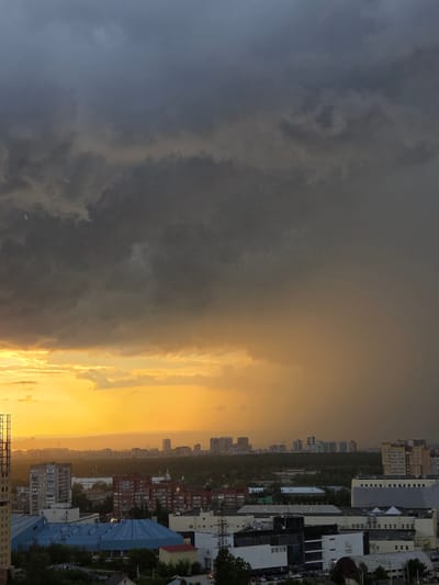 The image shows a city skyline at dusk with a dramatic sky filled with dark storm clouds. A bright band of sunlight is visible on the horizon, casting a golden glow over the scene. In the foreground, there are various buildings, including residential high-rises, industrial structures, and what appears to be a sports complex with a blue roof. The overall impression is one of impending weather change over an urban environment. The location is identified as Shirkovo, Russia.