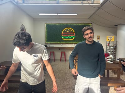 The image portrays an indoor scene within a casual dining establishment in Porto Salvo, Portugal. Two adult males are central subjects. The male on the left, with dark curly hair, wears a light-colored short-sleeved t-shirt with a "LEVI'S Stay Fresh" logo and dark trousers. He is positioned with his head tilted downwards, looking away from the viewer, and his arms are slightly bent with open hands at his sides. The male on the right, with dark hair and a mustache, wears a dark teal long-sleeved crew-neck sweater and light-colored cargo shorts. He is looking towards the left, with his left hand partially in his pocket.

The background features a large, rectangular wall-mounted sign displaying a neon outline of a hamburger (yellow bun, orange patty, green lettuce/sauce) against a green artificial grass-like panel. Below this sign, a wooden counter is equipped with two red stools. To the right of the counter, a metal shelving unit holds multiple white dishes and other items. A yellow sign attached to the shelving unit reads "OBRIGADO PELA VISITA!" (Thank you for your visit!). Another abstract decorative element is visible on the left wall. The room has light-colored walls, overhead fluorescent lighting, and a skylight is visible in the upper portion of the frame. Multiple wooden tables are scattered throughout the space, and the floor appears to be paved or tiled. A hand holding a smartphone is partially visible in the bottom right foreground, indicating the presence of another individual.