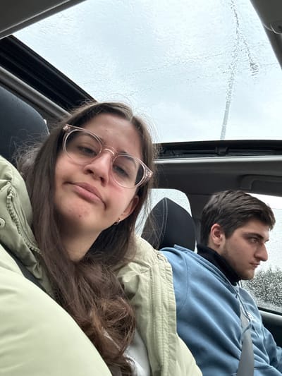 The image displays two individuals inside a vehicle, stated to be located in Zahesi, Georgia. In the foreground, a female with long brown hair and clear-framed eyeglasses occupies the passenger seat, wearing a light, possibly quilted, jacket and a small silver hoop earring on her left ear. She looks directly towards the camera with a neutral expression. Behind her and to the right, a male sits in the driver's seat, wearing a dark turtleneck under a light blue hoodie, secured by a seatbelt. He is observed looking out the right-side window, away from the camera. The vehicle features an open glass sunroof and side windows, all exhibiting visible raindrops, indicating current or recent precipitation. Through the sunroof, an overcast sky is visible, and through the side window, blurred green foliage and an indistinct dark vertical object are present in the background.