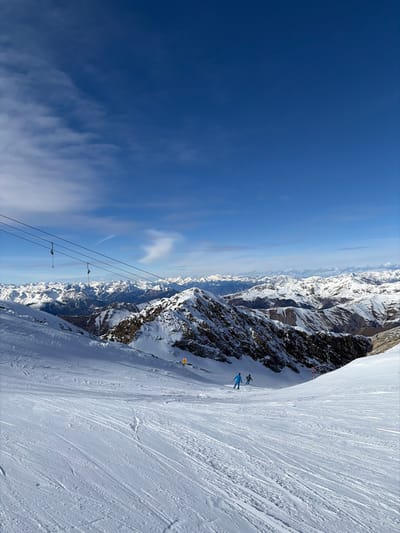 The image displays a high-alpine ski slope within the mountainous region of Tux, Austria. The foreground and midground feature a wide, groomed piste covered in snow, marked by numerous ski tracks. Two individuals, identified as skiers, are actively descending the slope in the mid-ground. The lead skier, towards the left, wears a blue full-body ski suit. A second skier, in darker apparel, follows slightly behind and to the right.

In the upper left quadrant, a ski lift system with dark cables and several empty hangers extends diagonally across the frame. The environment includes rugged, snow-dusted rock formations flanking the groomed trail. A yellow rectangular marker is visible on the left side of the piste, with a red pole marker further down on the right. The background consists of expansive, snow-capped mountain ranges under a clear, deep blue sky, accented by wispy white clouds.