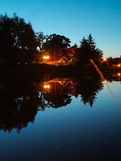 This image depicts a serene twilight scene by a body of water, likely in Rumia, Poland. In the background, a house with a red-tiled roof and illuminated windows is visible, surrounded by dark trees. Two bright street lamps cast a warm glow on the scene. The water in the foreground acts as a mirror, reflecting the house, trees, and lights with remarkable clarity, creating a symmetrical and tranquil ambiance. The sky is a deep, clear blue, transitioning from a lighter shade at the horizon to a darker hue overhead, characteristic of dusk. There are no people visible in the image. The overall atmosphere is peaceful and picturesque, highlighting the beauty of the natural and architectural elements at twilight.