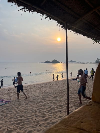This image depicts a beach scene at sunset in Gokarna, India, viewed from underneath a thatched roof structure supported by a vertical wooden pole on the right. The sky shows a hazy gradient from pale orange around the low-hanging sun on the upper right to light grey further up. The sun's light creates a shimmering golden reflection across the calm ocean water, where small waves are breaking near the shore. Several dark, rocky islets are visible offshore.

The beach itself is composed of light brown sand with visible ripples and depressions, indicative of foot traffic. In the immediate foreground, a shirtless male with dark hair, wearing dark shorts, walks barefoot towards the right. Another shirtless, bearded male in dark shorts, carrying a clear plastic bottle in his left hand, walks barefoot towards the right, partially obscured by the wooden pole and a foreground barrier on the right.

Across the mid-ground, numerous individuals are engaged in various activities. On the left side of the beach, a group of three males interacts near the waterline; one appears to be holding a mobile phone in a manner consistent with taking a photograph. Further left, another individual walks along the shore. Near the center, several people are wading or swimming in the ocean, and a red buoy or float is visible. On the right side of the beach, a group of people, mostly silhouetted, walk along the shoreline. One person is identifiable by a yellow vest or jacket. Further back on the sand, a small group appears to be playing a ball game, and another individual is lying down. A colorful patterned textile is laid on the sand in the lower-left foreground.