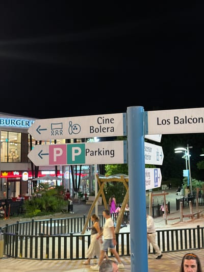 The image shows a street scene at night, likely in Alcorcón, Spain, with several signs directing visitors. The main subjects are the signs themselves, which indicate "Los Balcones," "cine Bolera," and "Parking." The "Parking" signs are color-coded, with one green and one pink, and both have an arrow pointing downwards. There are also icons on the signs suggesting a bowling alley and perhaps restrooms or other facilities.
In the background, there are people visible, some walking and others seated at tables, suggesting a public area with businesses or entertainment venues. The lighting indicates it is nighttime, with artificial lights illuminating the scene. The overall setting appears to be an outdoor commercial or entertainment district. The presence of people, the lighting, and the directional signs suggest a lively atmosphere. One person is partially visible in the upper right corner, looking towards the camera with a smile. The scene conveys a sense of activity and accessibility for visitors.