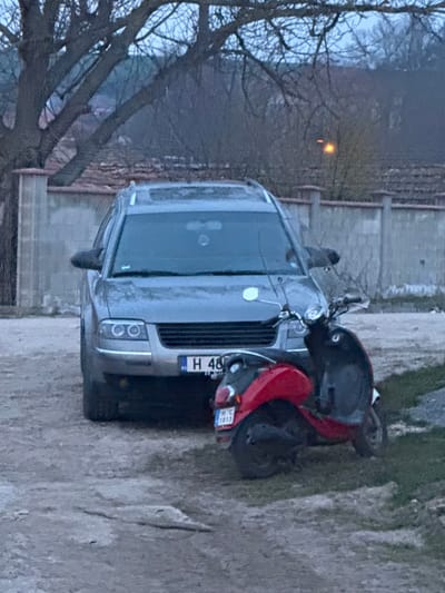 The scene depicts an unpaved outdoor area in Omarchevo, Bulgaria, under subdued lighting. A gray Volkswagen Passat B5.5 station wagon is parked facing the viewer. The vehicle features circular headlights, roof rails, and a sunroof, with a partially visible Bulgarian front license plate reading "H 48XX XX". Positioned in front and slightly to the right of the car is a red and black scooter, also stationary. Its rear license plate, clearly visible, displays "H C 1013". The immediate ground consists of dirt with sparse patches of grass. In the mid-ground, a concrete wall with reddish-brown coping stones spans horizontally. Beyond this wall, the background includes bare deciduous trees and distant terrain. A single, bright orange light source is visible in the upper right background. No individuals are present in the image, and no actions or interactions are depicted.