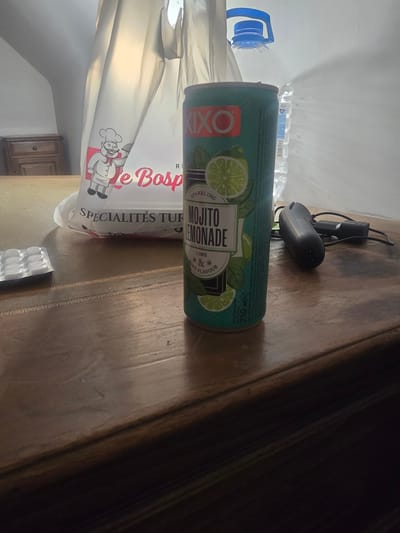 A close-up, eye-level shot shows a wooden table with various items on it. To the right, a green can of "XIXO MOJITO LEMONADE" stands prominently. Next to it, a black computer mouse with its wire and a power adapter are visible. In the background, a large white plastic bag with a chef illustration and "Le Bosp..." text is partially visible. Behind the bag, a large clear plastic bottle with a blue cap can be seen. To the left of the can, a blister pack of white circular tablets is on the table. The environment appears to be an indoor setting with a dresser visible in the far background. The image does not provide any specific information or context about the city of Saint-Nazaire, France.