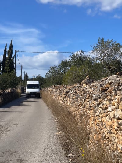 An outdoor scene depicts a narrow asphalt road in la Riera de Gaià, Spain, under a partially cloudy blue sky. A white panel van, possibly a Fiat Ducato, with license plate "XXXX CMJ" on the front, is driving towards the viewer in the center of the road. A single individual is visible in the driver's seat. The road is flanked on both sides by dry-stacked stone walls composed of irregular light brown and tan rocks. Tall, dry brown grasses and weeds line the immediate edges of the asphalt next to these walls, with a few small yellow flowers visible on the right. Beyond the walls, verdant trees and dense foliage are present. On the left side of the road, several utility poles with overhead lines run parallel to the road. In the background on the left, several tall, conical evergreen trees, possibly cypresses, stand prominently. The scene is illuminated by direct sunlight, casting subtle shadows.