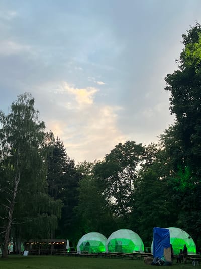 The image depicts an outdoor gathering at dusk within the Sremski Karlovci Municipality, Serbia. The scene is characterized by a partially cloudy sky with shades of light blue, grey, and hints of yellow-orange from the setting sun, visible towards the upper-right portion of the frame. A dense, dark green treeline forms the background, comprising various deciduous and coniferous trees.
In the midground, a flat grassy area features multiple temporary structures. Four white geodesic domes are prominently displayed, each internally illuminated with a vibrant green light. Inside these domes, tables and benches are arranged, and the silhouettes of several individuals are visible. Between the second and third domes from the left, a large blue tarp is draped over an unseen structure, with a small hay bale and dark bags or equipment positioned at its base. Approximately 5-7 individuals are gathered around this blue tarp and the rightmost dome; some are seated on benches or the hay bale, while others stand.
To the left of the domes, a wooden pergola or shelter is adorned with multiple strings of warm white Edison-bulb-style lights. Underneath this pergola, several wooden picnic tables and benches are set up. A white patio umbrella stands open nearby, alongside two white rectangular signs or boards leaning against a support. On the far left of the image, a tall tree with light-colored, somewhat bare branches stands out against the darker background foliage. The foreground consists of a mowed grassy field, populated with additional wooden picnic tables and benches arranged for seating. The overall lighting suggests late evening, with artificial illumination from the green domes and string lights complementing the fading natural light.