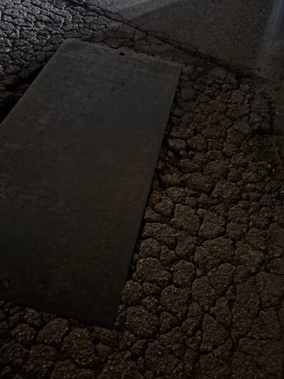 The image shows a rectangular metal plate resting on a heavily cracked asphalt surface. The asphalt exhibits a pattern of irregular, polygonal segments, suggesting advanced deterioration. The scene appears to be dimly lit, with strong contrast and deep shadows obscuring finer details. It is unknown if this image was taken in Quart de Poblet, Spain, but nothing in the image suggests this as fact. The plate's surface is rough and dark, reflecting ambient light in a mottled fashion. There are no people, subjects, actions, or interactions visible. The environment is limited to the immediate area of the plate and the asphalt.