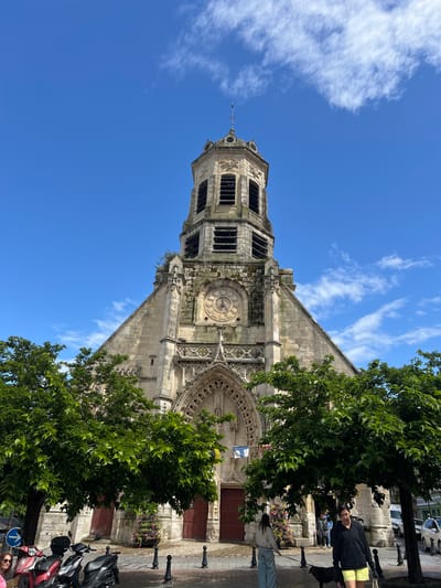 An eye-level, medium shot captures the imposing facade of the Église Sainte-Catherine, a historic church in Honfleur, France, under a bright blue sky dotted with wispy white clouds. The church, constructed from light-colored stone, features a prominent central bell tower with arched openings and a cross atop its octagonal lantern. Decorative stonework adorns the facade, including a large circular clock and intricate gothic-style arches framing the main entrance. Two large, dark red wooden doors lead into the building. In the foreground, lush green trees with dense foliage partially obscure the lower portion of the church. In front of the church, parked on a cobblestone street, are two red and black scooters. Several people are visible near the entrance of the church, including a woman in a black hoodie and yellow shorts walking a black dog, and another person in a grey sweatshirt and blue jeans walking with a leash. The street is lined with black bollards. The overall impression is a sunny day in a historic European town.