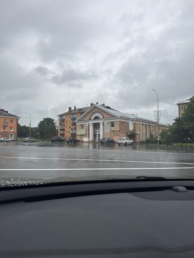 The image depicts a street scene in Kondopoga, Russia, on a cloudy, overcast day, possibly during or just after rainfall, as evidenced by the wet pavement and visible water droplets on the window in the foreground. The orientation of the image is portrait, with the street running vertically from bottom to top.
The scene features several buildings lining the street, with architectural styles varying from older, classical facades with decorative elements like columns and arched windows to more modern, multi-story apartment buildings. One prominent building in the mid-ground appears to be a public or commercial establishment, with signage visible that reads "КАФЕ" (Cafe). Several cars are parked along the roadside or driving on the wet street, including a white hatchback, a dark sedan, and a silver sedan. Trees with green foliage are visible between buildings and along the street, suggesting it is either spring or summer.
Streetlights with decorative lamps are present, along with power lines. The sky is heavily clouded with a uniform grey tone, indicating a lack of direct sunlight. The overall mood conveyed by the weather and the wet conditions is somber or subdued. The image appears to be taken from inside a vehicle, looking out through a side window, as indicated by the dark, textured panel on the right side of the frame, possibly part of the car's interior.