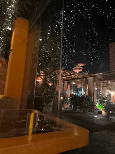 The image presents a serene nighttime view of an outdoor dining or lounge area, likely part of a restaurant or cafe, observed from an interior space through a large, reflective glass pane. In the immediate foreground, a corner of a polished wooden table with a dark, reflective surface is visible, showing a yellow object, possibly a pen, reflected on it.

Beyond the glass, the scene is illuminated by an array of warm, inviting lights. Prominent architectural features include large, orange-brown pillars and structures, along with pergolas featuring latticework ceilings. Several elegant, tiered lanterns with warm orange glows hang throughout the space, suspended from structures and within trees. The outdoor area is rich with dark foliage and numerous potted plants and bushes of various sizes, some of which are subtly lit from below with green or white accent lights.

Countless small white fairy lights are strung through the trees and foliage, creating a dazzling, starry effect that is further amplified by their reflections on the glass panel, blurring the distinction between the actual outdoor space and the reflections. In the far background, faint blue and white lights suggest additional structures or the wider urban environment.

The overall atmosphere is tranquil and sophisticated, suggesting a peaceful evening. No people are visible in the scene. The warm lighting, earthy tones, and architectural style evoke a luxurious Mediterranean or resort-like ambiance, consistent with a setting like Palaio Faliro, Greece. There is no visible text.
