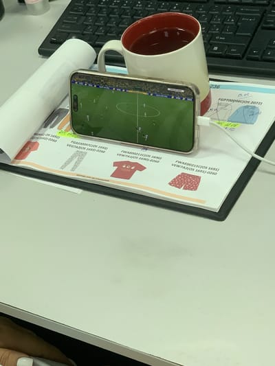This is a close-up, rotated shot of a desk scene. The main focus is a smartphone displaying a soccer game, propped up by a white coffee mug. The phone's screen shows a bright green soccer field with small figures of players in action. Along the bottom edge of the phone screen, there's a narrow band with "VISA" repeated multiple times. The mug is white with a red interior and has a charging cable plugged into the phone.
The background consists of papers and a keyboard. One paper has a series of alphanumeric codes and some text in a foreign language, possibly Russian ("повтор-пис не отшиваем"). There are also some diagrams and what appear to be images of clothing items on the papers. A black keyboard is visible on the left side of the image.
The scene suggests a casual moment at a desk, possibly during a workday or a study session, where the person is taking a break to watch a soccer match. The lighting is typical of an indoor environment. There are no people clearly visible, but a hand with white-painted fingernails is present on the right edge of the frame, suggesting someone is nearby, possibly holding or operating the phone or the camera. The visible text includes codes like "FGPT00098C(OS 2072)" and "FWAE00211C(OS 1690)," along with dates and times like "13.06." and "Ans 102.06" and "18.00". The presence of "VISA" on the phone screen and the international codes on the papers hint at a global context for the items on the desk. There are no strong indicators of time of day or weather.