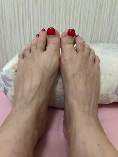 A top-down, close-up, eye-level shot shows two bare feet, with red nail polish on the toenails, resting on a pink surface and a pillow with a floral pattern. The background is a textured, vertical white curtain. There are no people or animals visible. The feet appear to be clean and well-maintained, with visible veins and a scar on the top of the left foot. The lighting is soft and even, casting no strong shadows. The scene is indoors and appears to be a bedroom or similar domestic setting. There is no information in the image to indicate it is located in Kortrijk, Belgium.