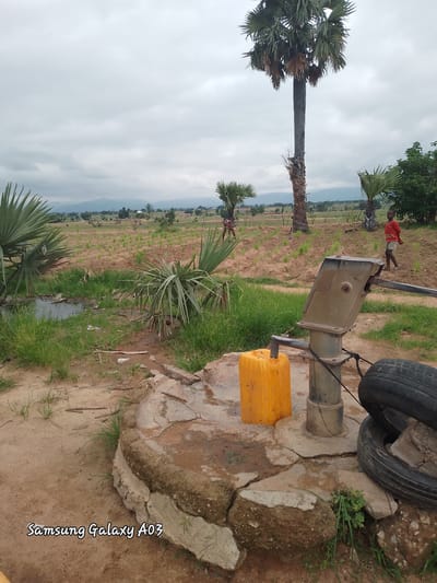 The image depicts an outdoor scene at a water pump in Angwan Hausawa, Nigeria. In the foreground, a manual water pump stands on a cracked concrete base, with a yellow jerrycan positioned to its left and two car tires to its right. Behind the pump, a young boy in a red shirt and shorts stands, looking towards the right. Further in the background, palm trees and other vegetation are visible, with fields and scattered buildings suggesting a rural or semi-rural settlement. The sky is overcast. The presence of the water pump and the jerrycan indicates water collection activities. The overall impression is of a functional, utilitarian setting within a community.